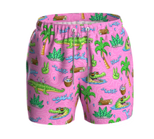 Load image into Gallery viewer, Pink swim shorts with tropical pattern featuring crocodiles, palm trees, and coconuts on a white background.