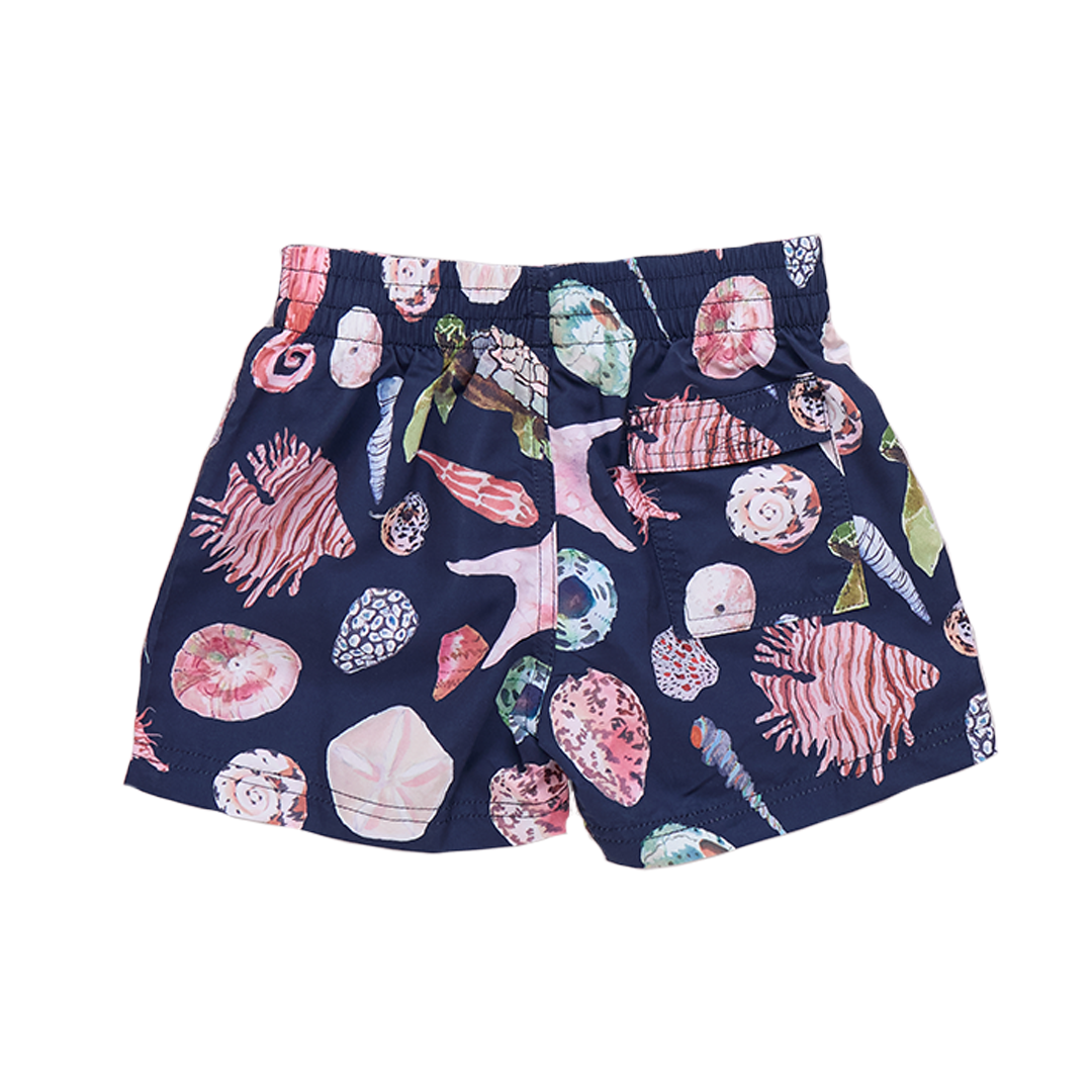 Baby boy in navy watercolor shell swim trunks by Pink Chicken, showing the colorful shell pattern on the back of the shorts.
