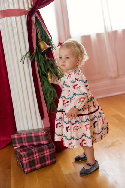 Baby girl wearing Pink Chicken Maribelle dress with Santa garland print, smiling and sitting on a cozy white surface.
